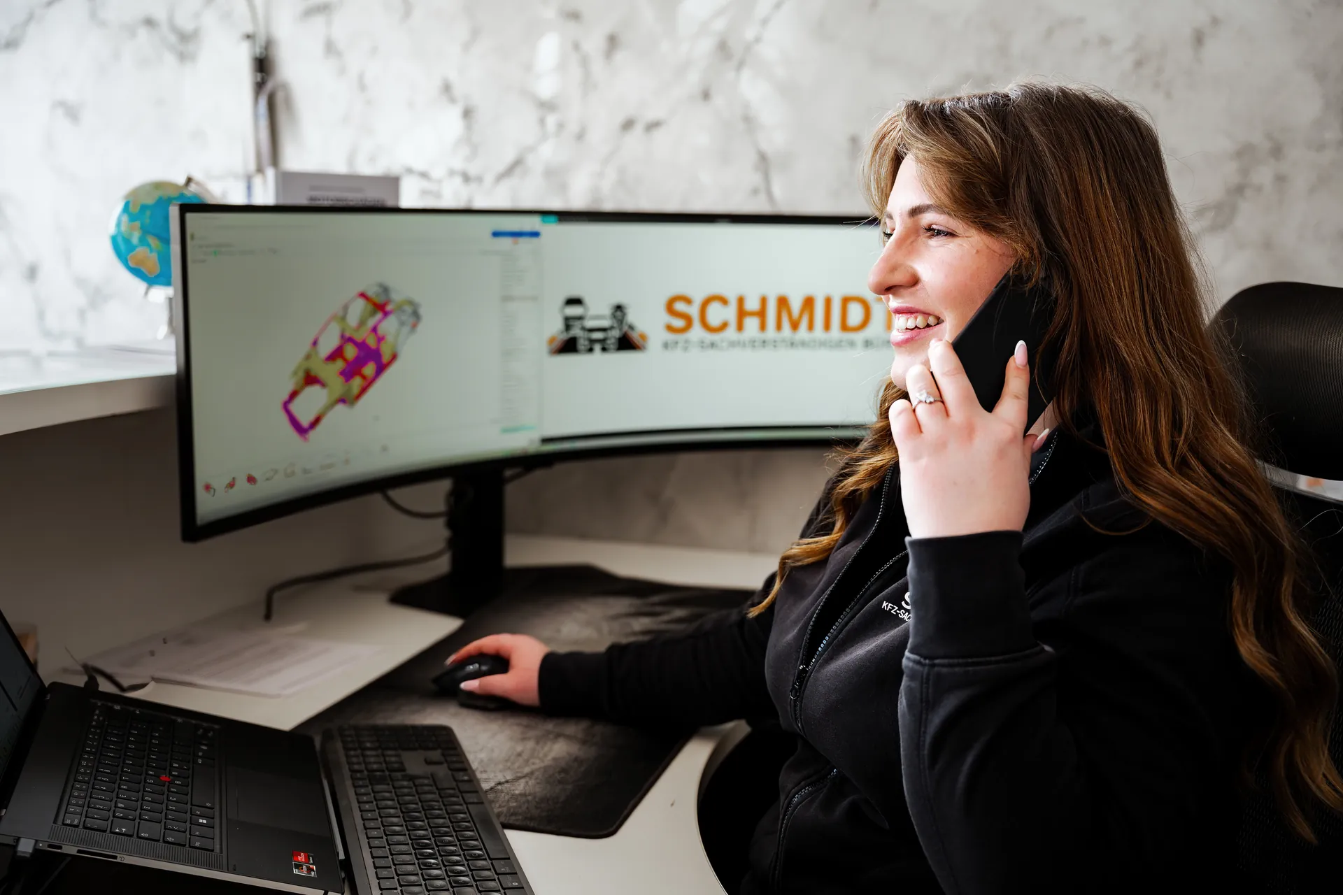 Mitarbeiterin des Sachverständigenbüros Schmidt telefoniert am Schreibtisch, im Hintergrund eine Karosserie-Schadensanalyse am Monitor